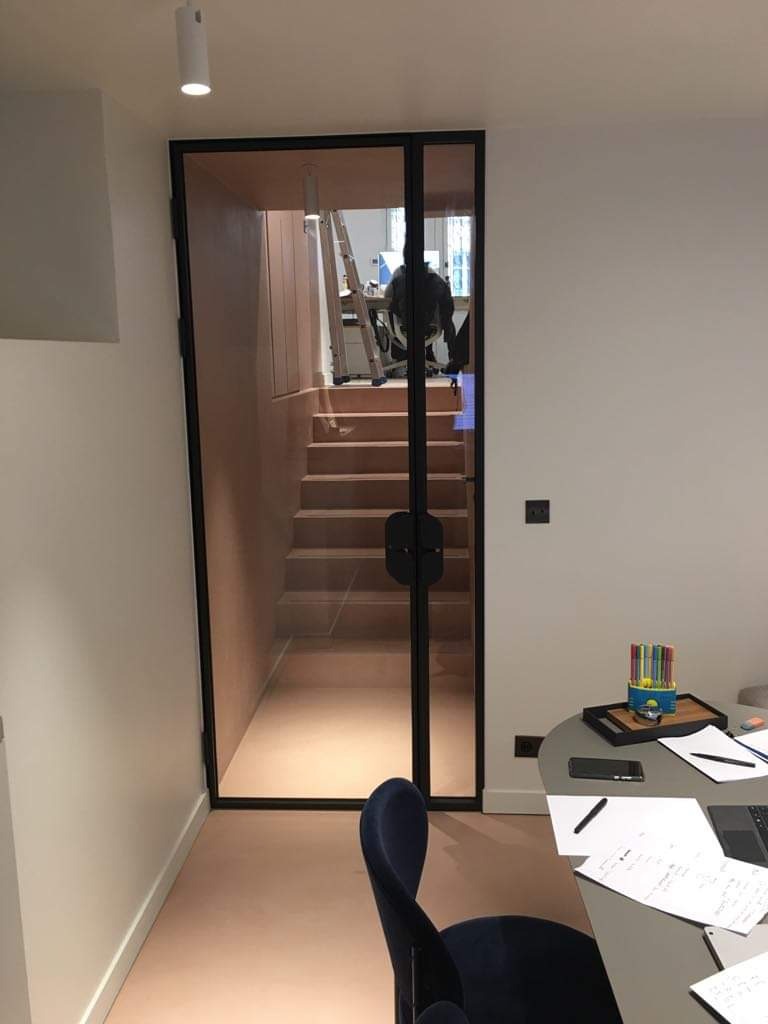 Szklane drzwi z czarną ramą prowadzące do klatki schodowej, widoczne biurko z przyborami i laptopem.