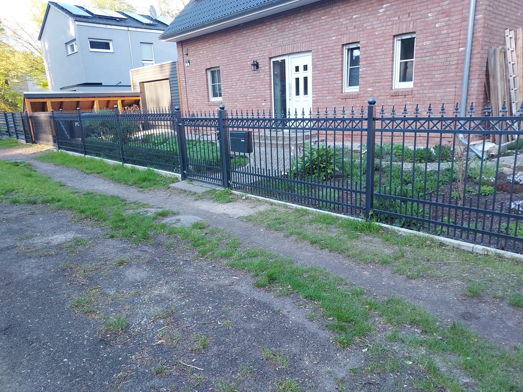 Ciemne, metalowe ogrodzenie z dekoracyjnymi detalami, oddzielające posesję z ceglanym domem i nowoczesnym budynkiem z panelami słonecznymi na dachu, widoczne z drogi z fragmentami trawy.