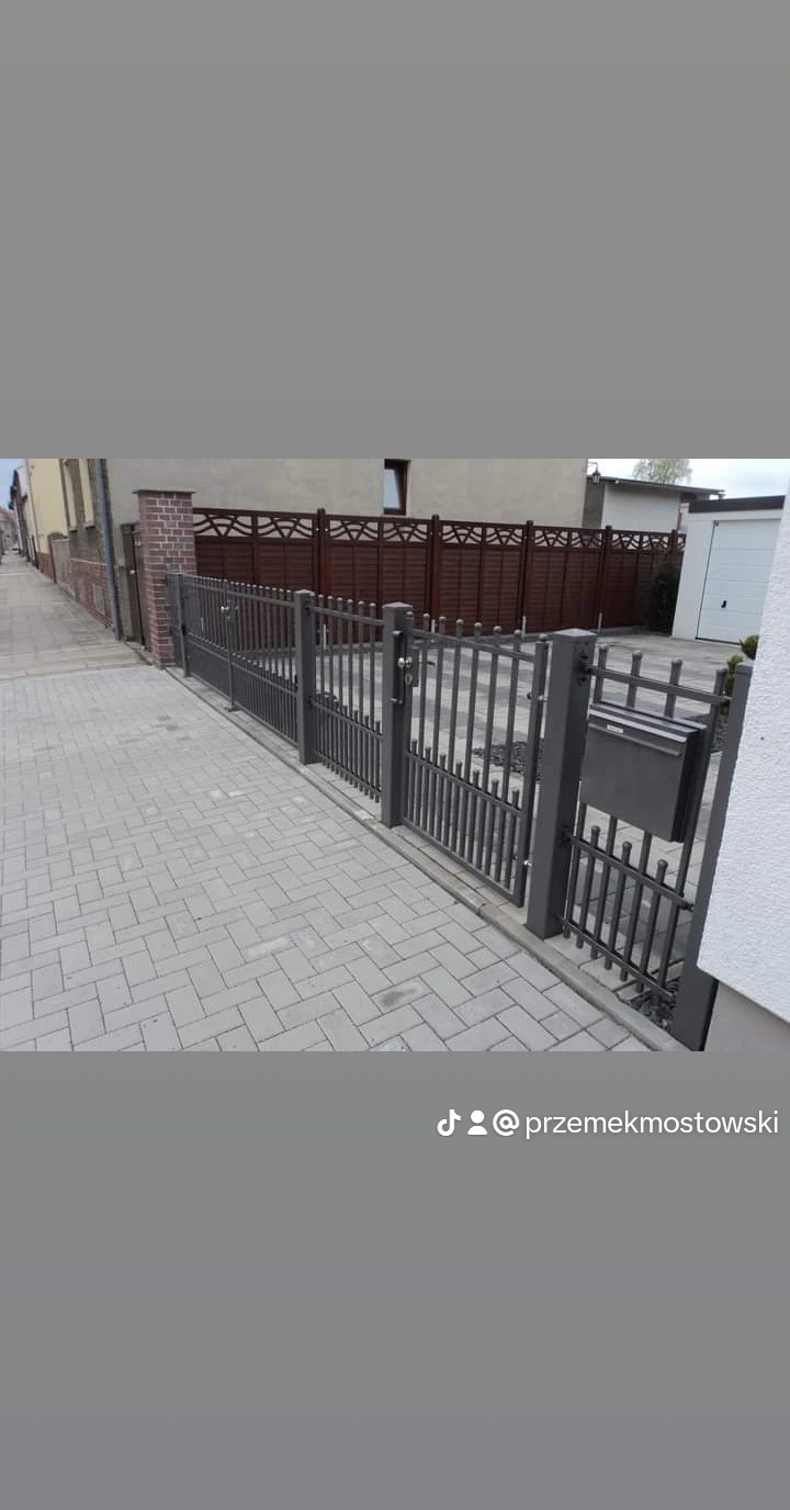 Szara metalowa brama wjazdowa z furtką i skrzynką na listy, widoczna z chodnika wyłożonego kostką brukową na tle domu i drewnianego ogrodzenia.