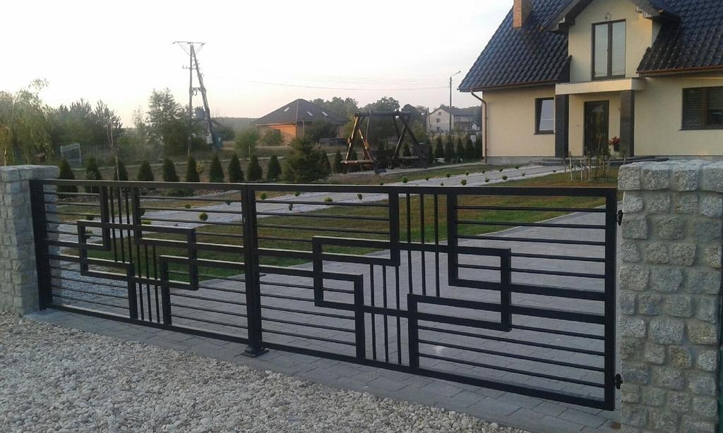 Nowoczesna, czarna brama wjazdowa z geometrycznym wzorem, osadzona na kamiennych słupkach, na tle domu z ciemnym dachem i zadbanego ogrodu.