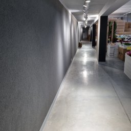 Pro-Tech - Długi korytarz w hurtowni kwiatów z szarą wykładziną na ścianie, oświetlony rzędem lamp sufitowych, z widocznymi kwiatami i roślinami w tle.