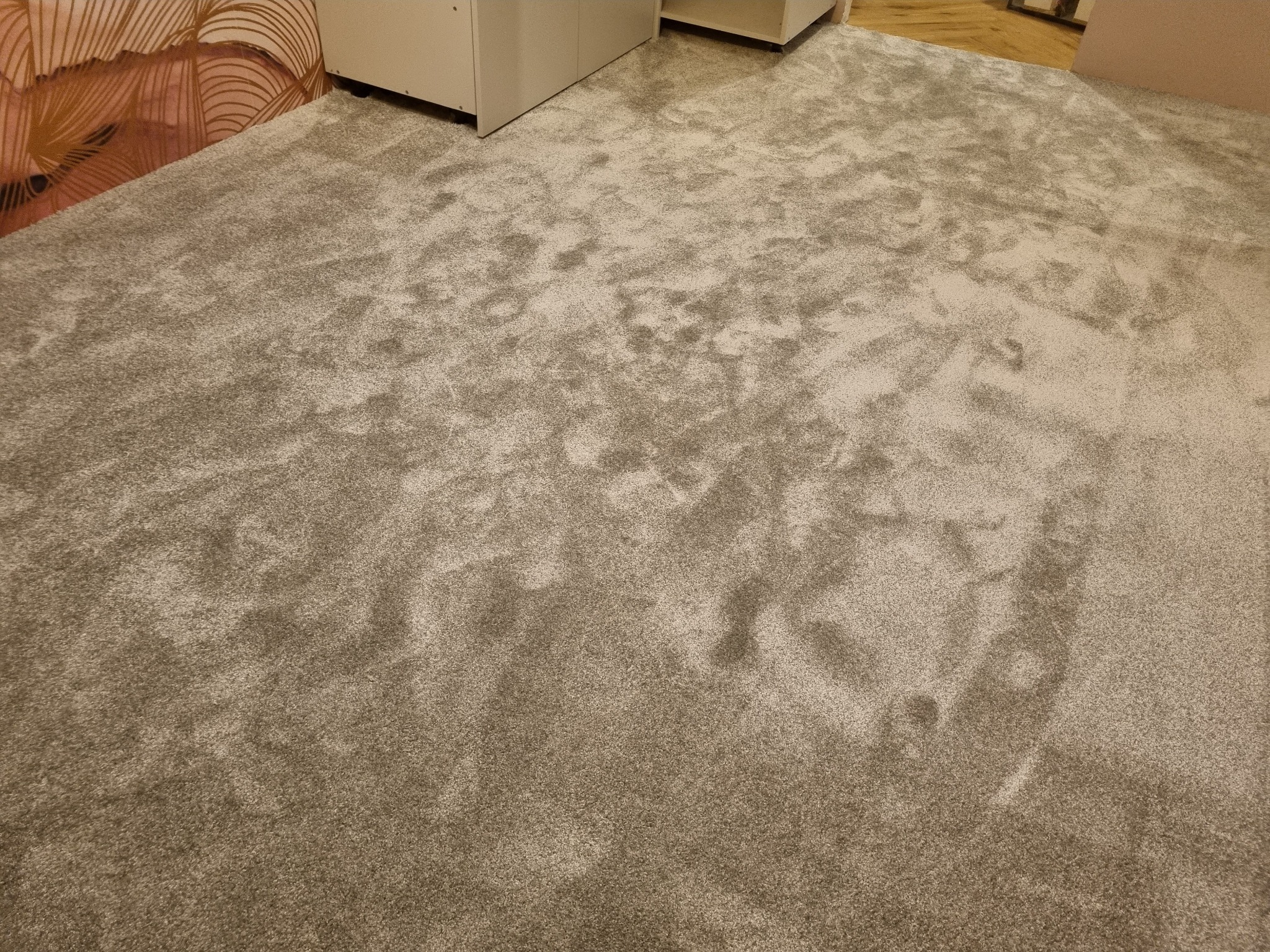 Szara, puszysta wykładzina dywanowa po montażu, widoczna przy ścianie z wzorzystą tapetą i białymi meblami.