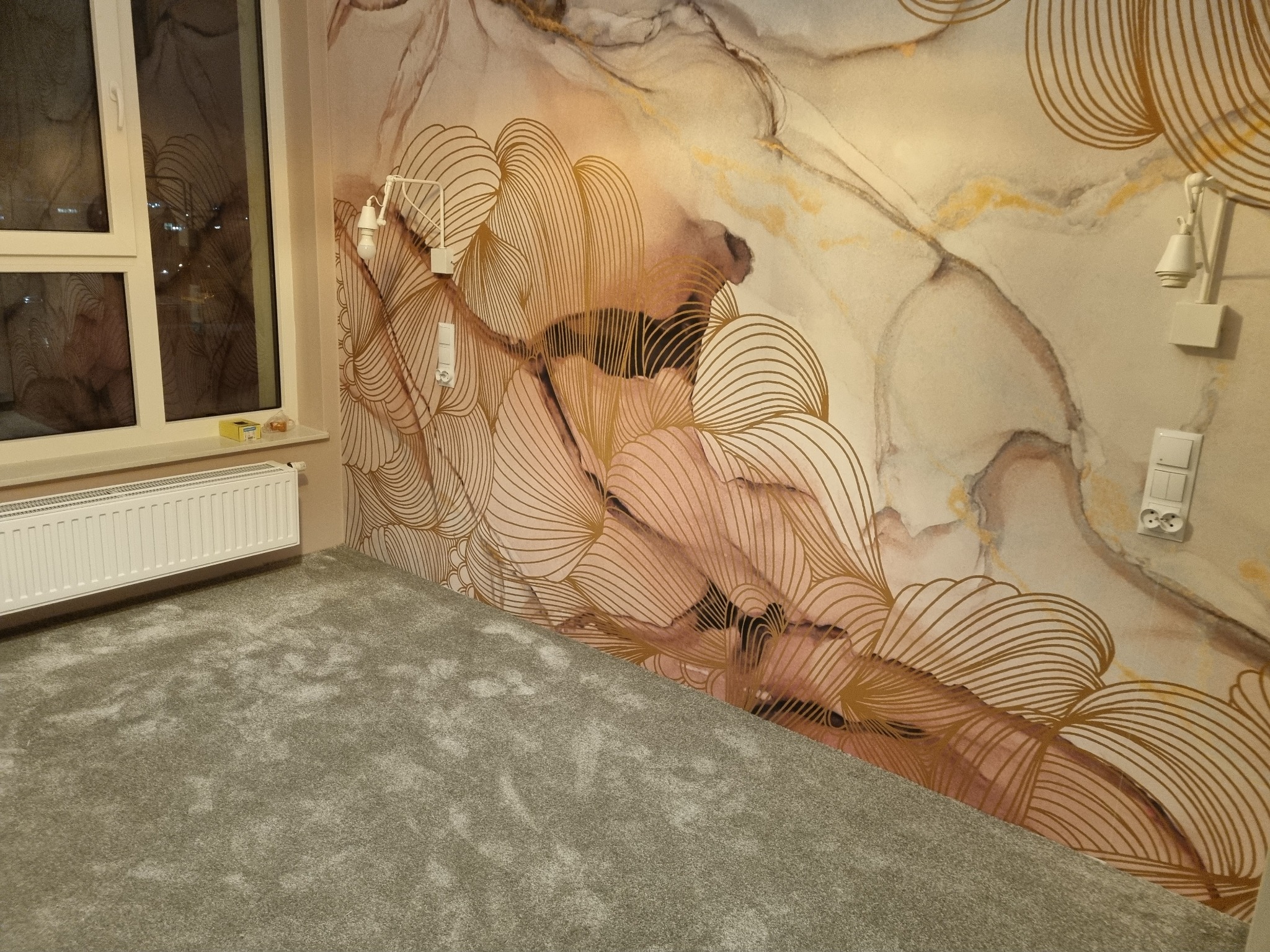 Jasnoszara wykładzina dywanowa w pokoju z grzejnikiem i ścianą pokrytą tapetą w abstrakcyjne, złote wzory.