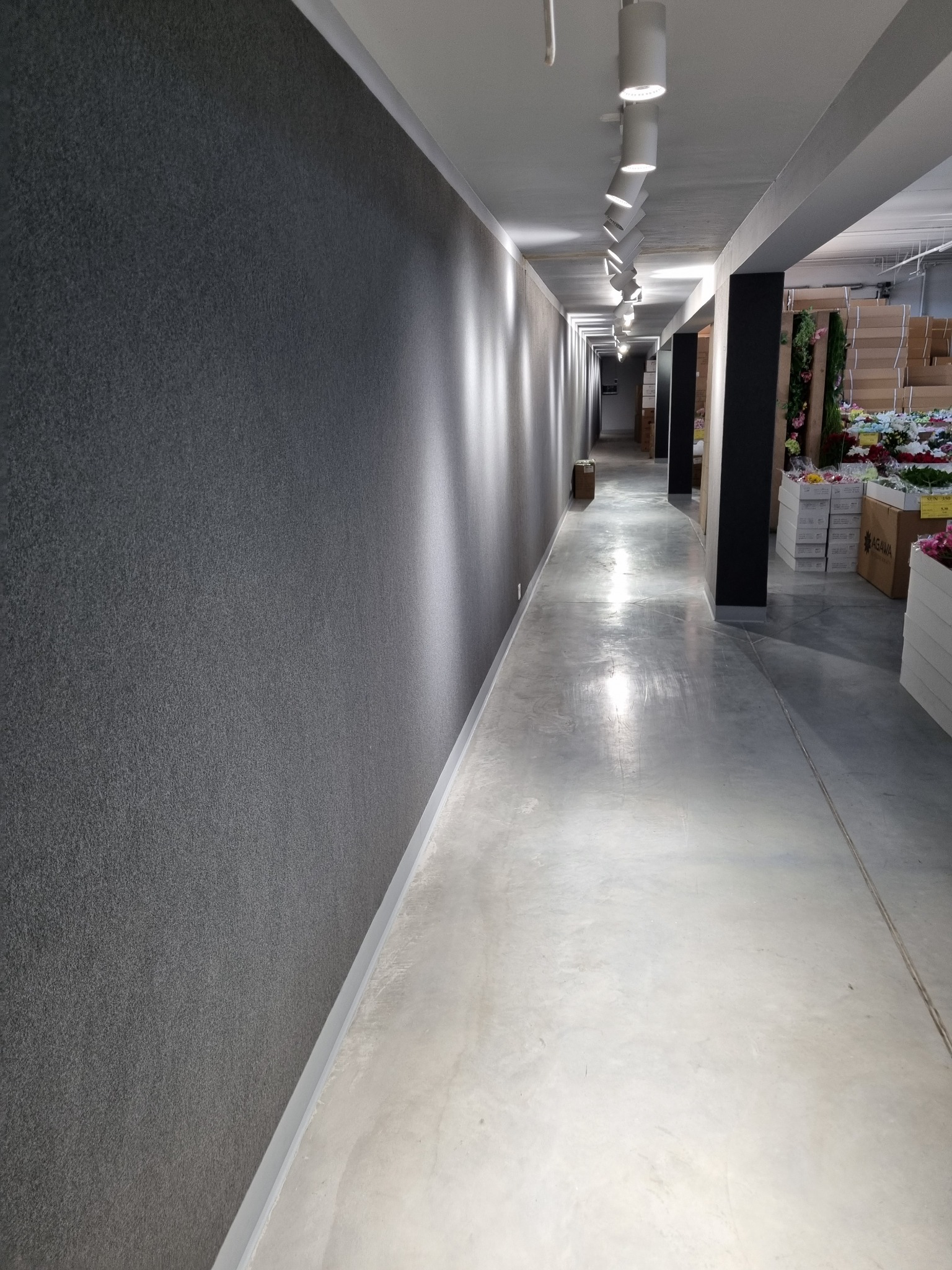 Długi korytarz w hurtowni kwiatów z szarą wykładziną na ścianie, oświetlony rzędem lamp sufitowych, z widocznymi kwiatami i roślinami w tle.