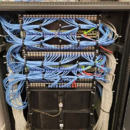 Serwerownia: szafa rack z przełącznikami sieciowymi i patch panelem, uporządkowane niebieskie kable Ethernet z czarnymi opaskami zaciskowymi, białe kable z tyłu, widoczne porty i diody LED.