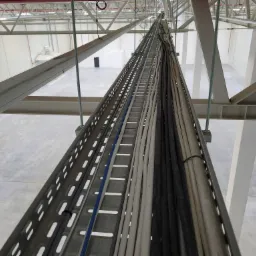 Metalowa korytka kablowa z ułożonymi przewodami, zawieszona pod sufitem w przestronnym, jasnym pomieszczeniu przemysłowym, widok z dołu.