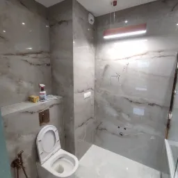 Nowoczesna łazienka w trakcie wykańczania, z marmurowymi płytkami, podwieszaną toaletą z bidetką w kolorze miedzi, minimalistycznym oświetleniem i widocznymi przewodami elektrycznymi.