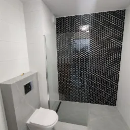 Wykończona łazienka z kabiną prysznicową i podwieszaną toaletą, ściana prysznica wyłożona czarną mozaiką heksagonalną, pozostałe ściany i podłoga w odcieniach szarości z imitacją marmuru.