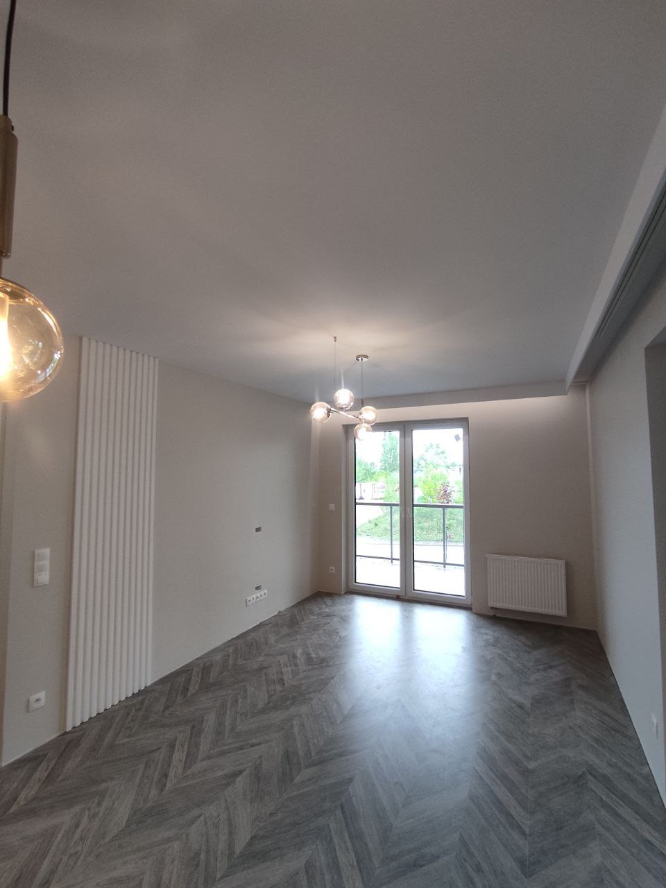 Jasne, przestronne wnętrze z parkietem ułożonym w jodełkę, dekoracyjnymi panelami ściennymi, nowoczesnym oświetleniem i balkonem z widokiem na zieleń.