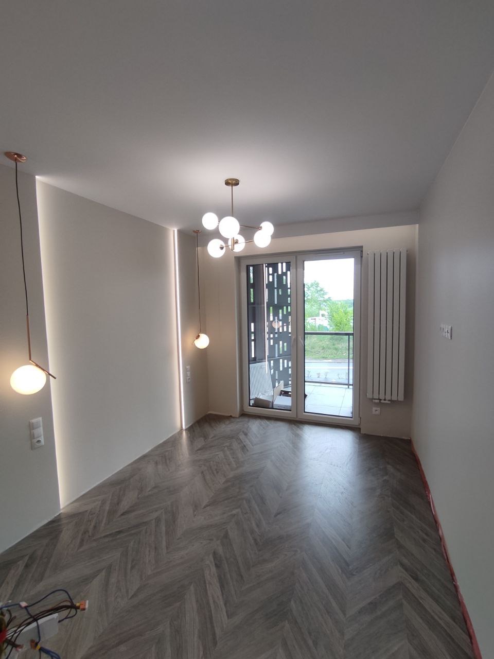 Nowoczesne wnętrze z ułożonym parkietem w jodełkę, oświetleniem punktowym i wiszącą lampą kulistą, widok na balkon przez przesuwne drzwi.