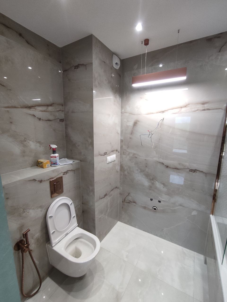 Nowoczesna łazienka w trakcie wykańczania, z marmurowymi płytkami, podwieszaną toaletą z bidetką w kolorze miedzi, minimalistycznym oświetleniem i widocznymi przewodami elektrycznymi.