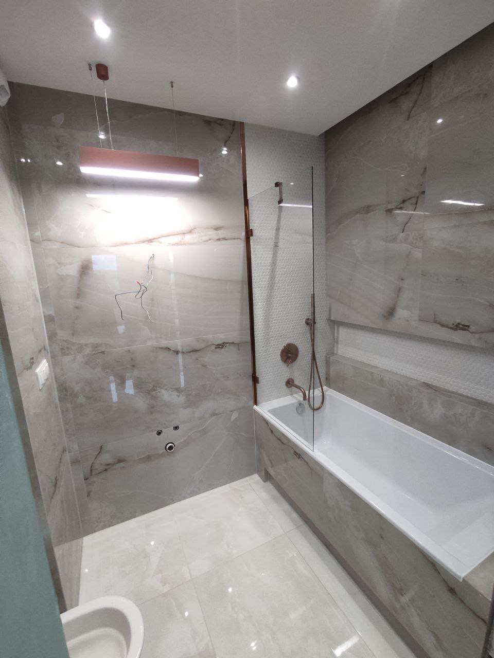 Wykończona łazienka z wanną obudowaną płytkami imitującymi marmur, szklanym parawanem nawannowym i armaturą w kolorze miedzi, podświetlona podłużną lampą sufitową.
