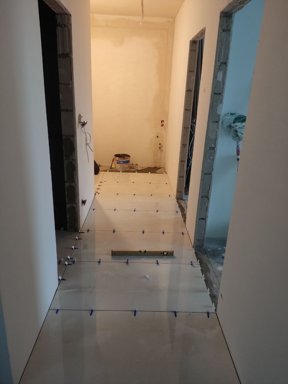Korytarz w trakcie układania jasnych płytek podłogowych z użyciem poziomicy i krzyżyków dystansowych; widoczne surowe ściany i przygotowane otwory drzwiowe.