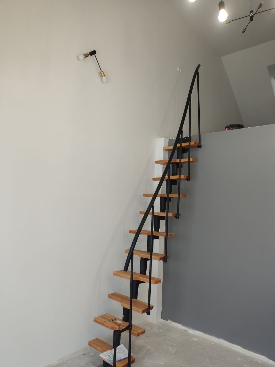 Wąskie schody spiralne z drewnianymi stopniami i czarną metalową konstrukcją, zamontowane w pomieszczeniu z szarą ścianą i białym sufitem, oświetlone lampami w stylu industrialnym.