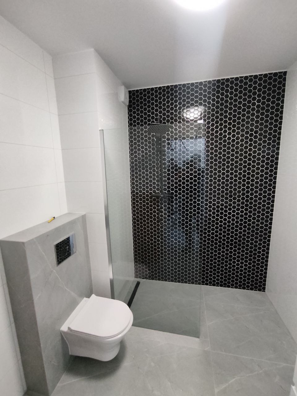 Wykończona łazienka z kabiną prysznicową i podwieszaną toaletą, ściana prysznica wyłożona czarną mozaiką heksagonalną, pozostałe ściany i podłoga w odcieniach szarości z imitacją marmuru.