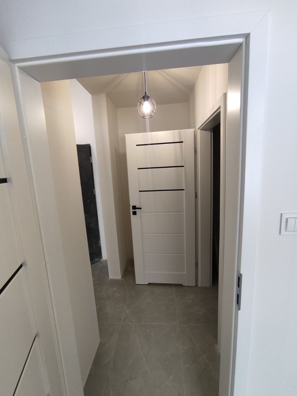 Korytarz po remoncie z szarą płytką podłogową, białymi drzwiami z czarnymi poziomymi paskami i ozdobną lampą wiszącą w metalowej klatce.