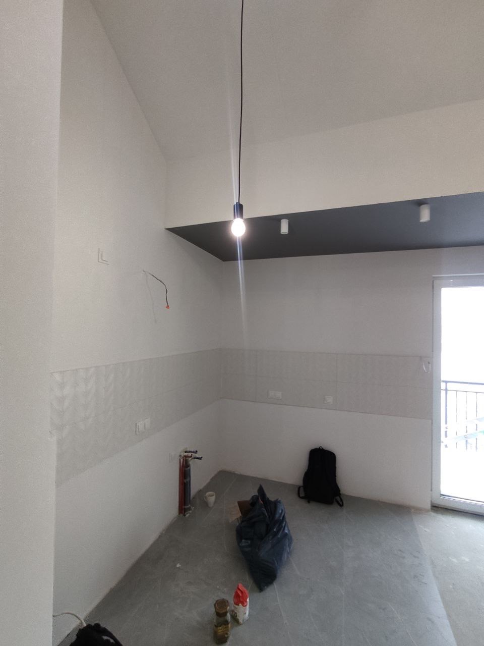 Wnętrze w trakcie wykańczania: ściany częściowo wyłożone płytkami, widoczne instalacje hydrauliczne, worki z materiałami budowlanymi, sufit z czarnym podwieszanym fragmentem i punktowym oświetleniem.
