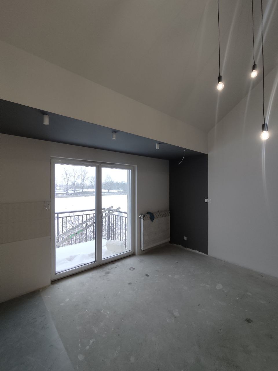 Surowe wnętrze pokoju z surowym betonowym wykończeniem podłogi, widokiem na zaśnieżony balkon i nowoczesnym oświetleniem wiszącym.