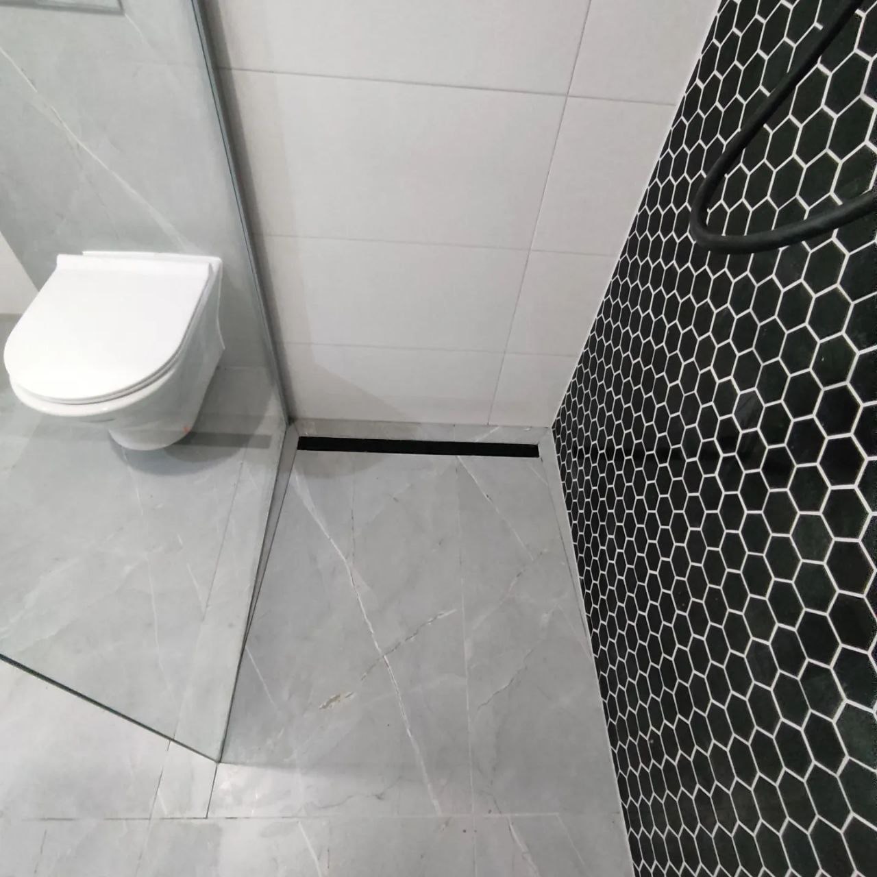 Nowoczesna łazienka w Katowicach z szarymi płytkami imitującymi marmur na podłodze i ścianach, kabina prysznicowa ze szkła oraz ściana wyłożona czarnymi heksagonalnymi płytkami.