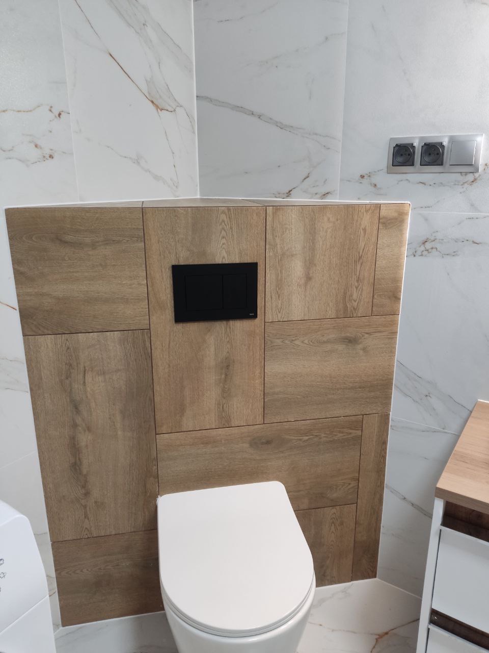 Nowoczesna toaleta z zabudową z płytek imitujących drewno, czarny panel sterujący spłuczką, marmurowe płytki na ścianach, gniazdka elektryczne w srebrnej ramce.