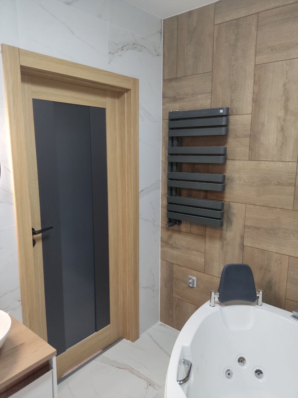 Nowoczesna łazienka z drewnopodobnymi płytkami na ścianie, grzejnikiem drabinkowym w kolorze grafitowym, białą wanną z zagłówkiem i drzwiami z szarą szybą.