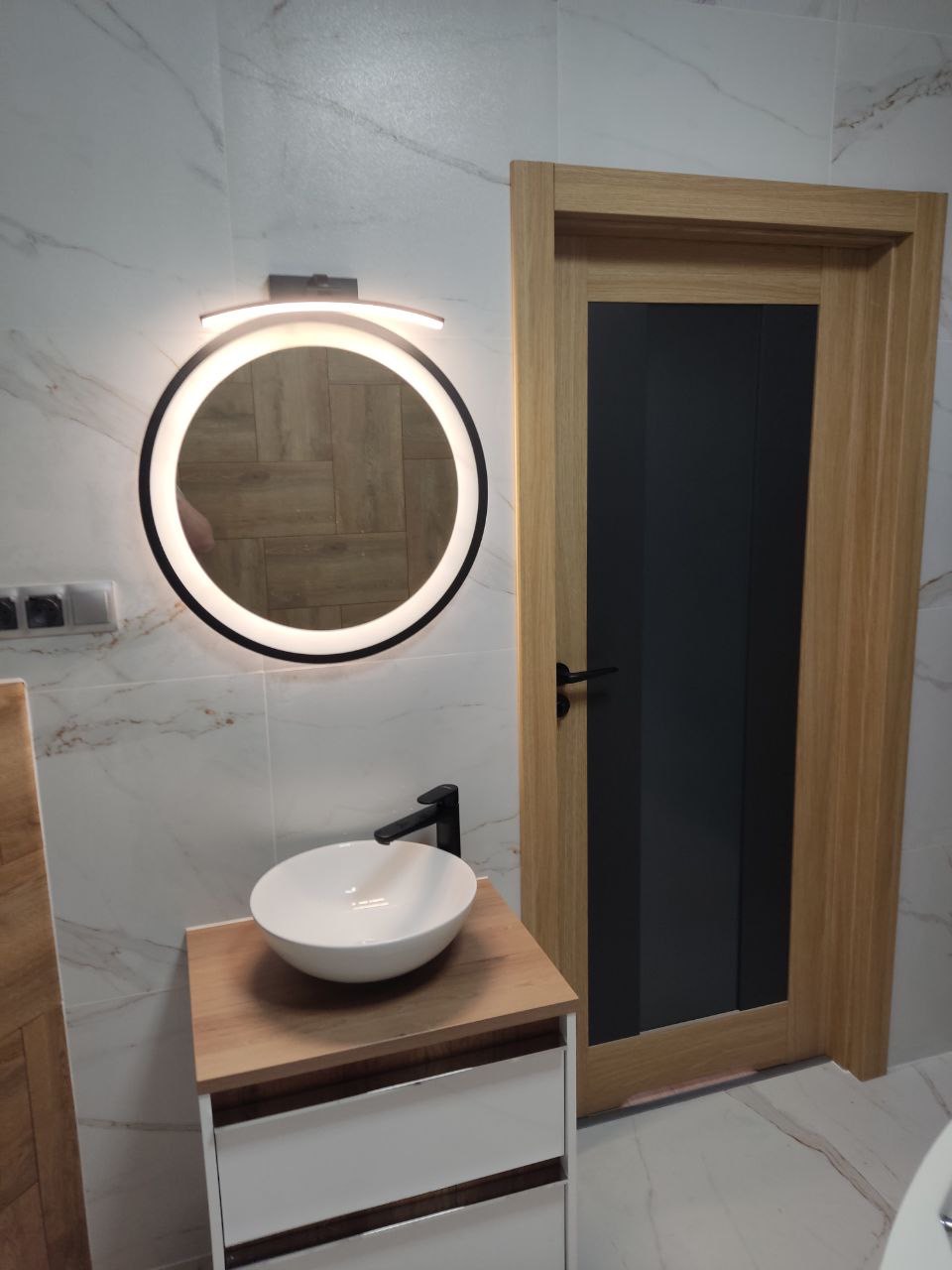 Nowoczesna łazienka z okrągłym lustrem podświetlanym LED, umywalką nablatową na szafce z białymi szufladami i drewnianym blatem oraz drewnianymi drzwiami z czarnym szkłem.