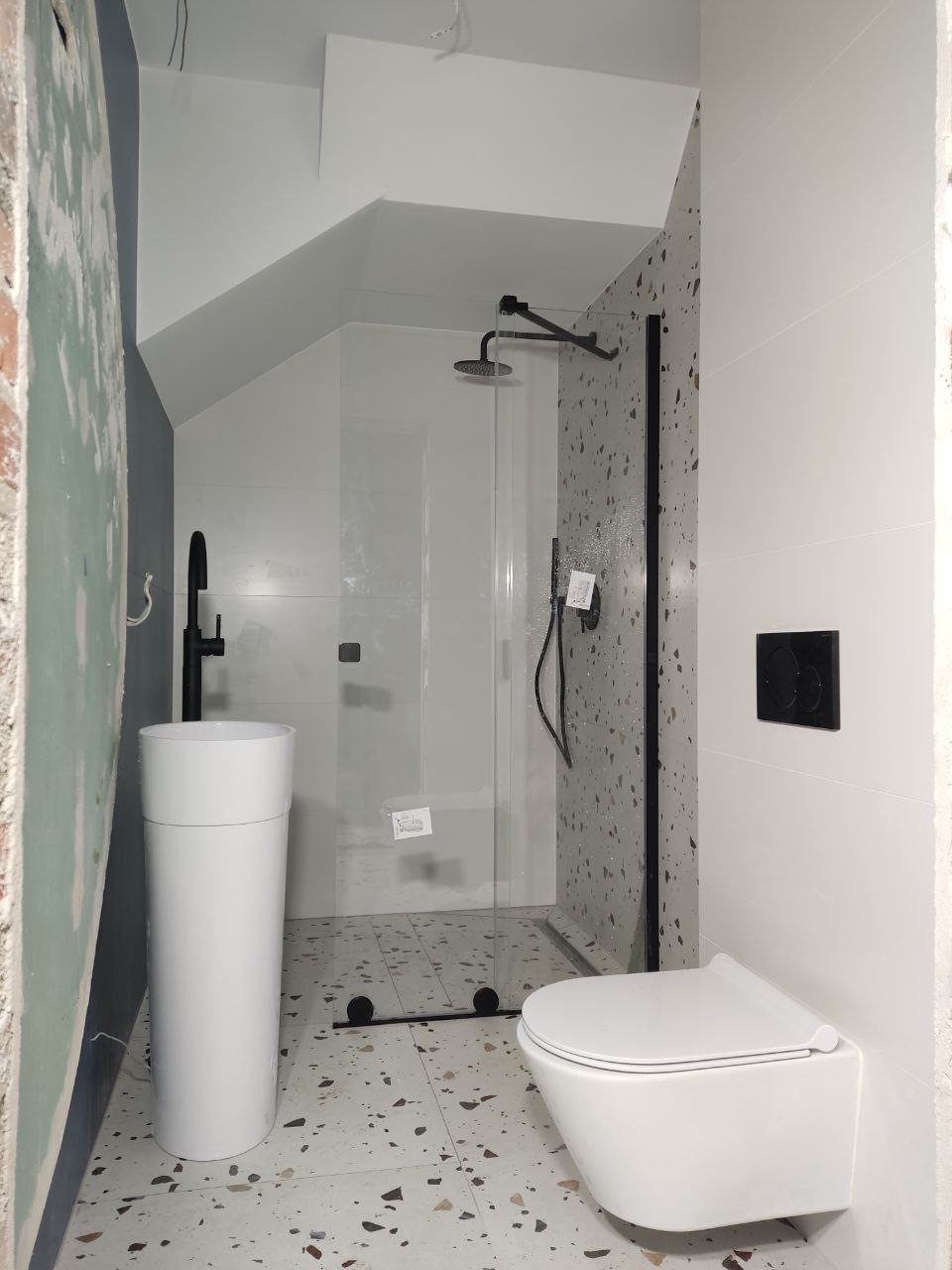 Nowoczesna łazienka w trakcie wykańczania: biała umywalka kolumnowa, podwieszana toaleta i szklana kabina prysznicowa z czarną armaturą na tle ściany w kropki i surowego muru.