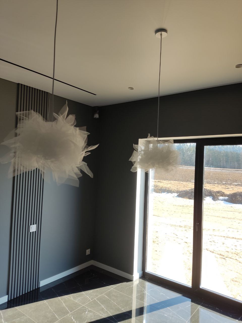 Szare wnętrze z ciemną podłogą, dekoracyjnymi lampami wiszącymi i pionowymi panelami ściennymi, widok na zewnątrz przez duże okno.