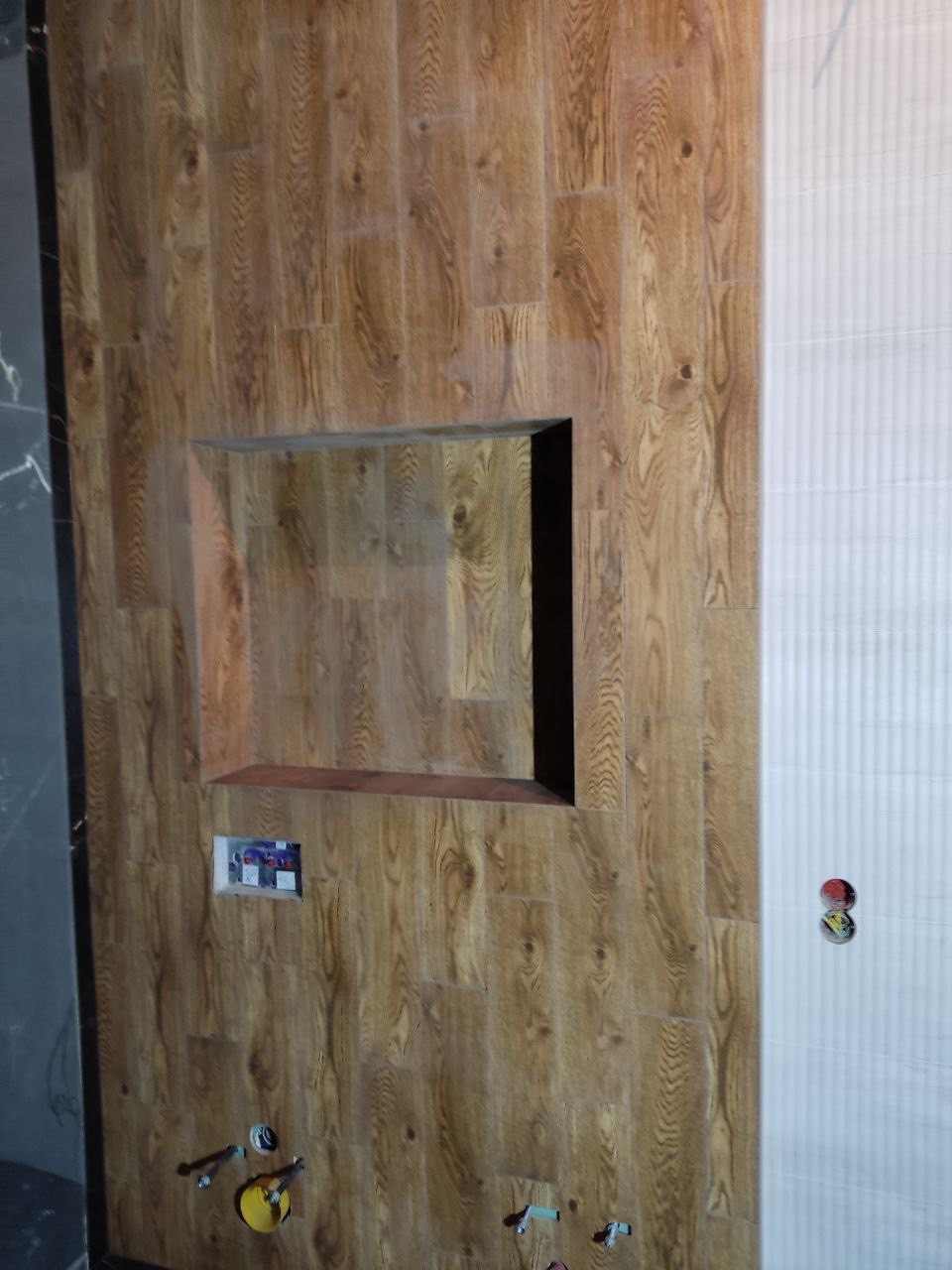 Ściana w łazience wykończona płytkami imitującymi drewno, z wnęką i przygotowanymi przyłączami hydraulicznymi, sąsiadująca z białą ścianą z delikatnym wzorem.