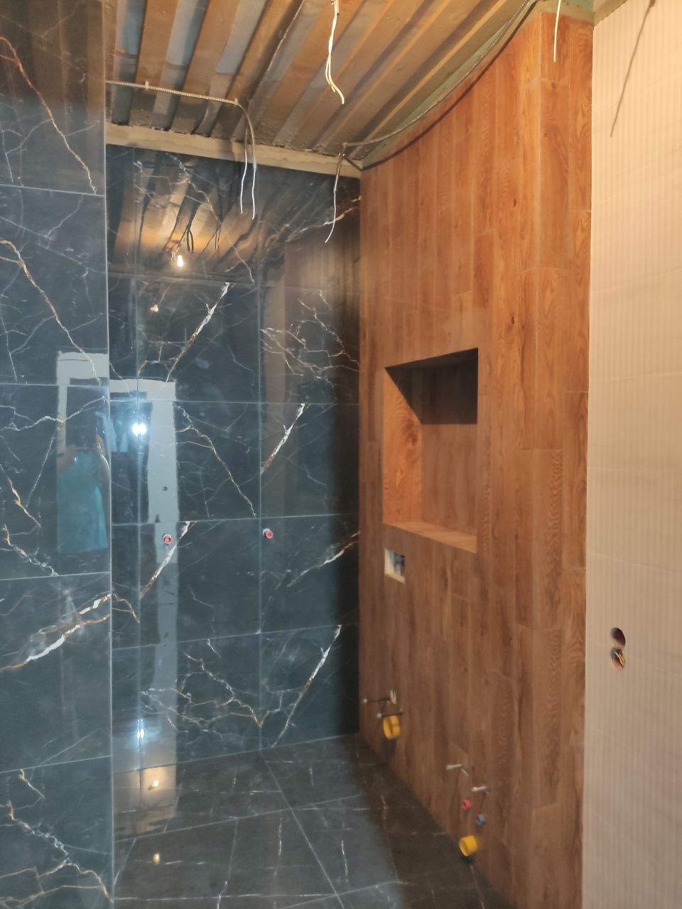 Wnętrze łazienki w trakcie remontu: ciemne płytki imitujące marmur z jasnymi żyłami na ścianach i podłodze, drewnopodobna obudowa z wnękami na ścianie, widoczne niedokończone instalacje i surowy...