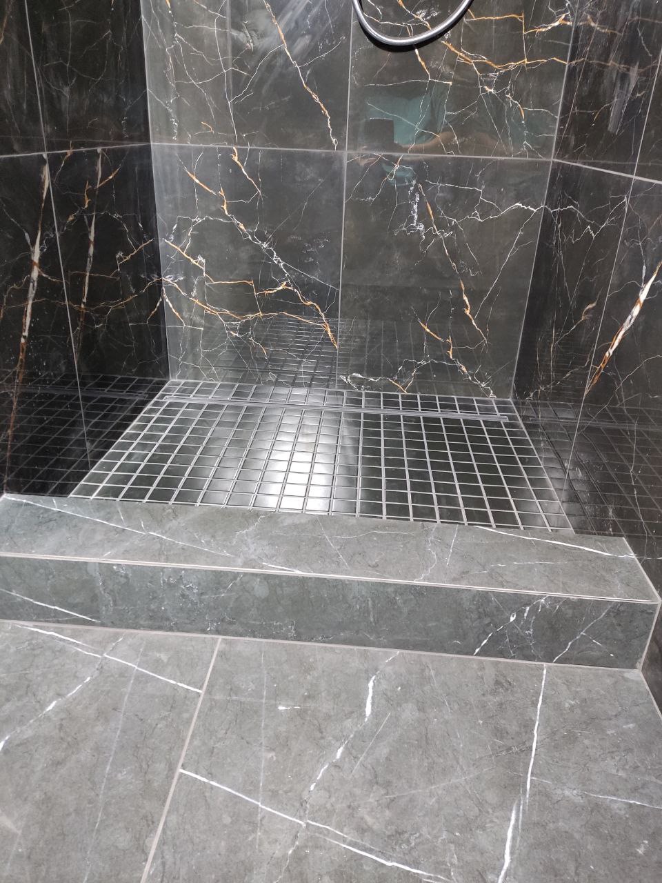 Wykończony brodzik z odpływem liniowym, wyłożony czarnymi płytkami imitującymi marmur z złotymi żyłami, oraz szarymi płytkami na podłodze i stopniu.