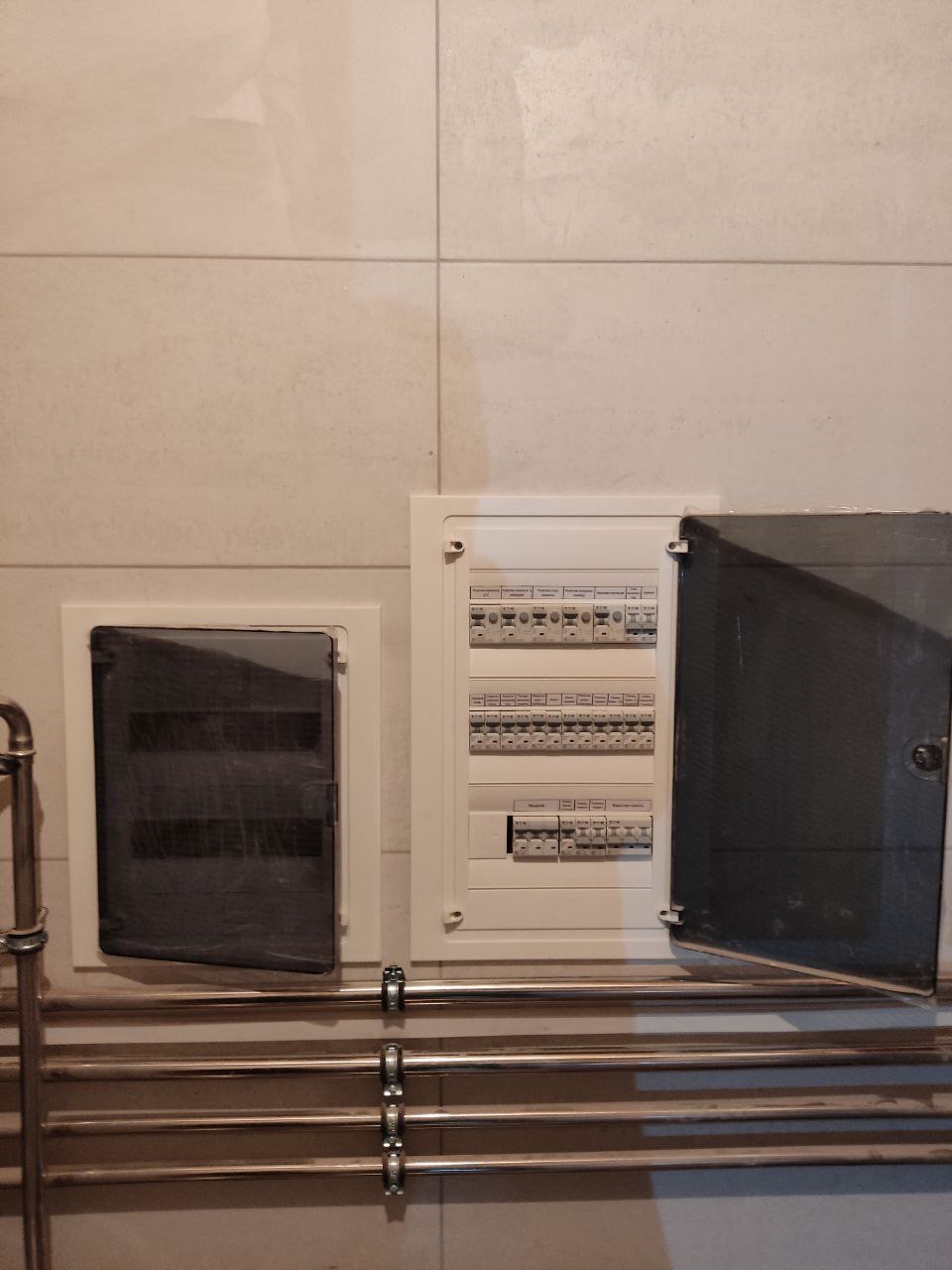 Dwie skrzynki elektryczne na kafelkowanej ścianie, jedna z otwartymi drzwiczkami, poniżej poziome rury grzejnika drabinkowego.