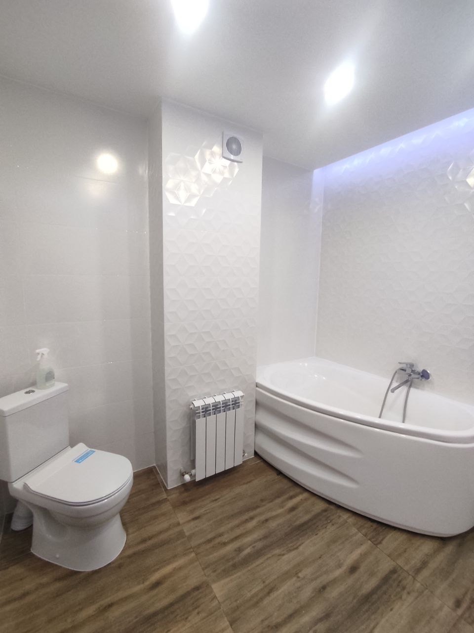 Nowoczesna łazienka z białą armaturą, grzejnikiem panelowym i drewnopodobną podłogą; ściana z dekoracyjną trójwymiarową fakturą, podświetlenie LED przy suficie.