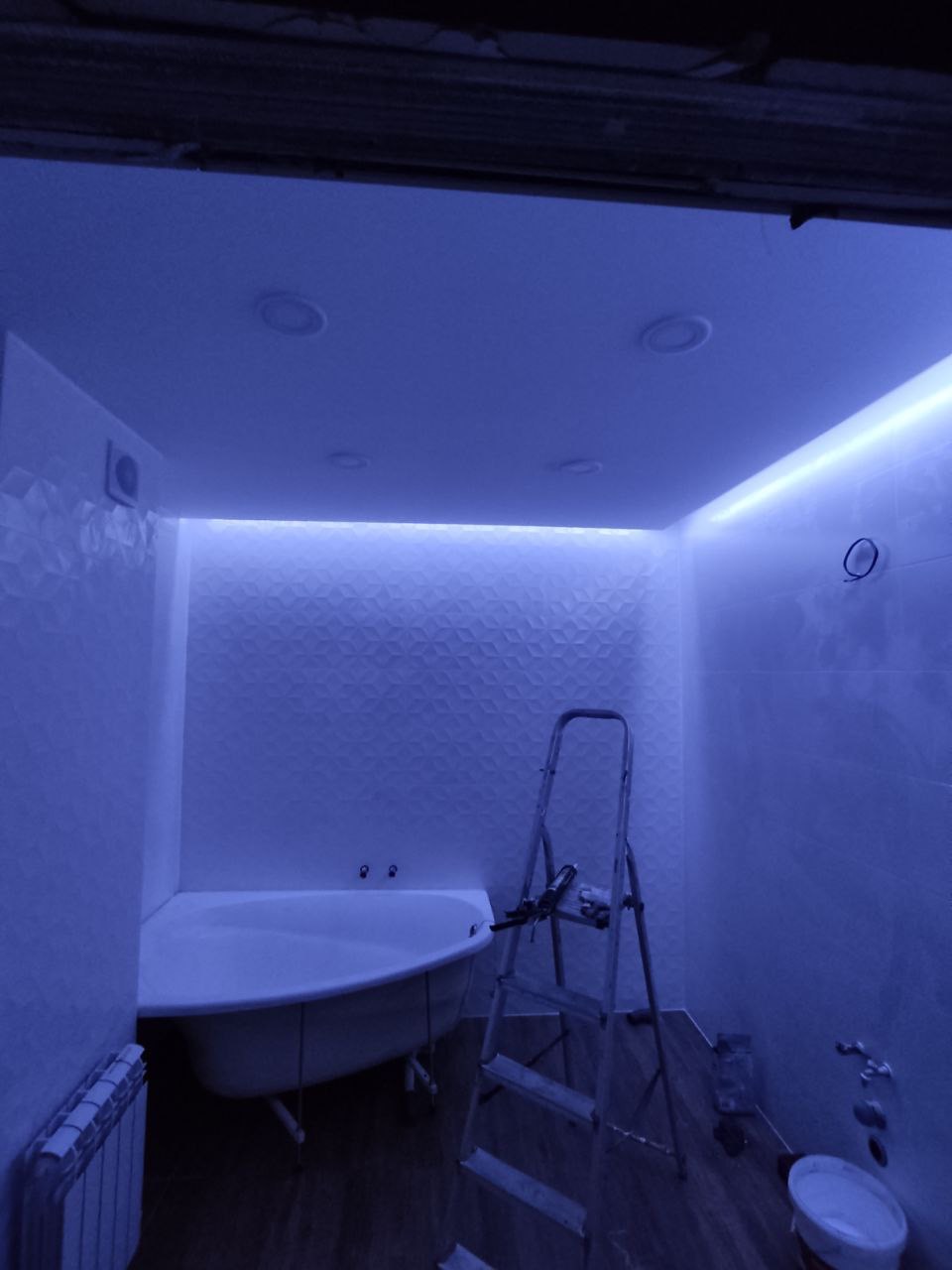 Niebieskie oświetlenie LED w łazience w trakcie remontu, widoczna wanna narożna, grzejnik, drabina i płytki 3D na ścianie.