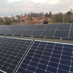 PROBO Bogusław Pabisz - Instalacja paneli słonecznych na dachu domu jednorodzinnego, widok z góry, pracownik w czarnej kurtce i czapce na dachu, w tle zabudowa mieszkalna i pochmurne niebo.