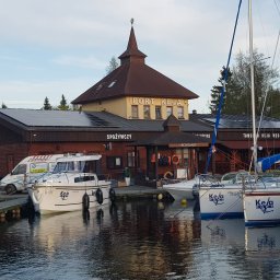 PROBO Bogusław Pabisz - Budynek portowy z panelami słonecznymi na dachu, widok z wody, kilka jachtów zacumowanych przy nabrzeżu.