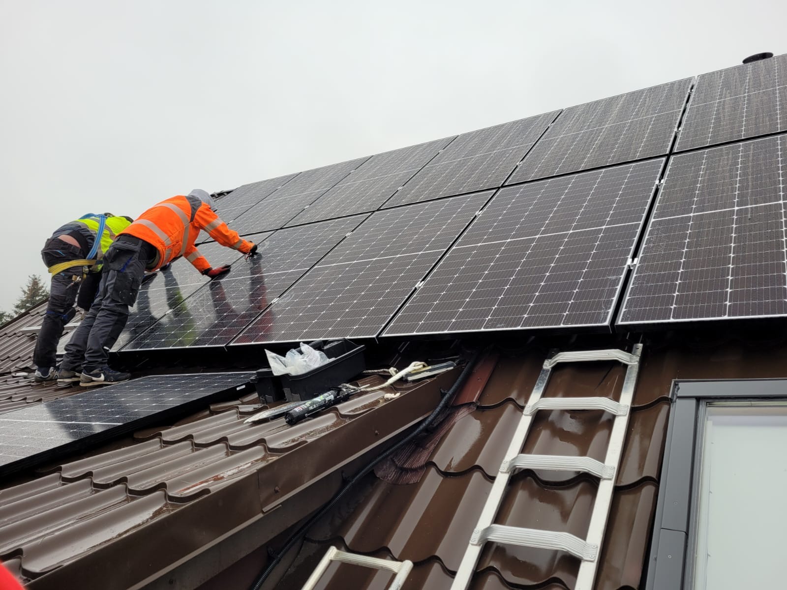 Montaż paneli słonecznych na brązowym dachu przez dwóch instalatorów w odblaskowych ubraniach, widoczna drabina i narzędzia.