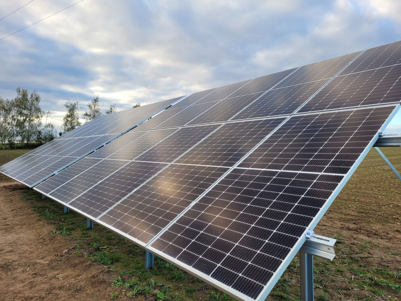 Rząd paneli słonecznych zamontowanych na metalowej konstrukcji na polu, z widocznym niebem z chmurami w tle.