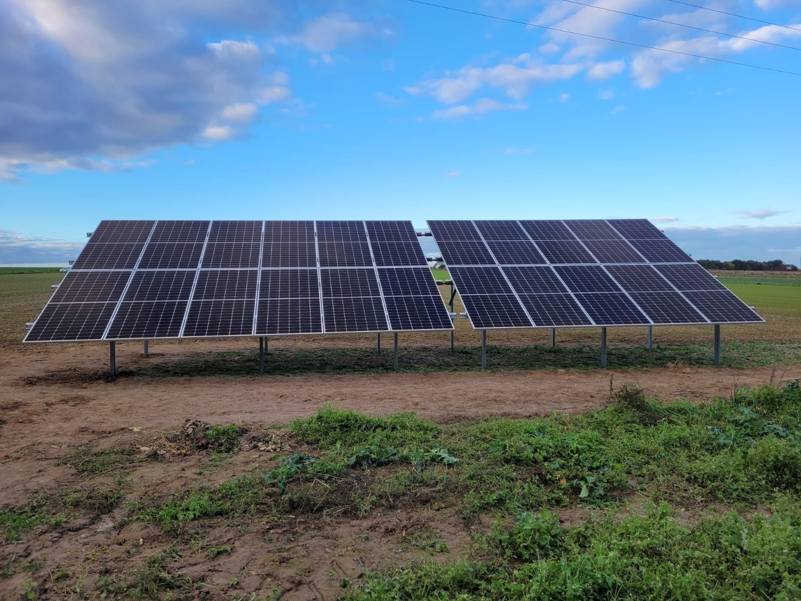 Dwa rzędy paneli fotowoltaicznych na metalowych konstrukcjach, ustawione na polu z widoczną linią energetyczną w tle, pod błękitnym niebem z chmurami.