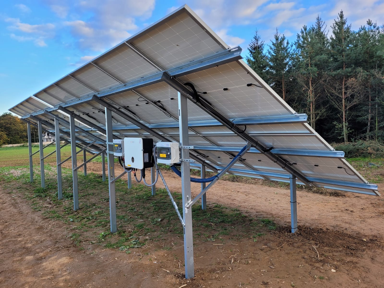 Widok od spodu na zainstalowane panele fotowoltaiczne z inwerterami, widoczne metalowe konstrukcje wsporcze, kable w niebieskich peszlach, tło to pole i las iglasty, pochmurne niebo.