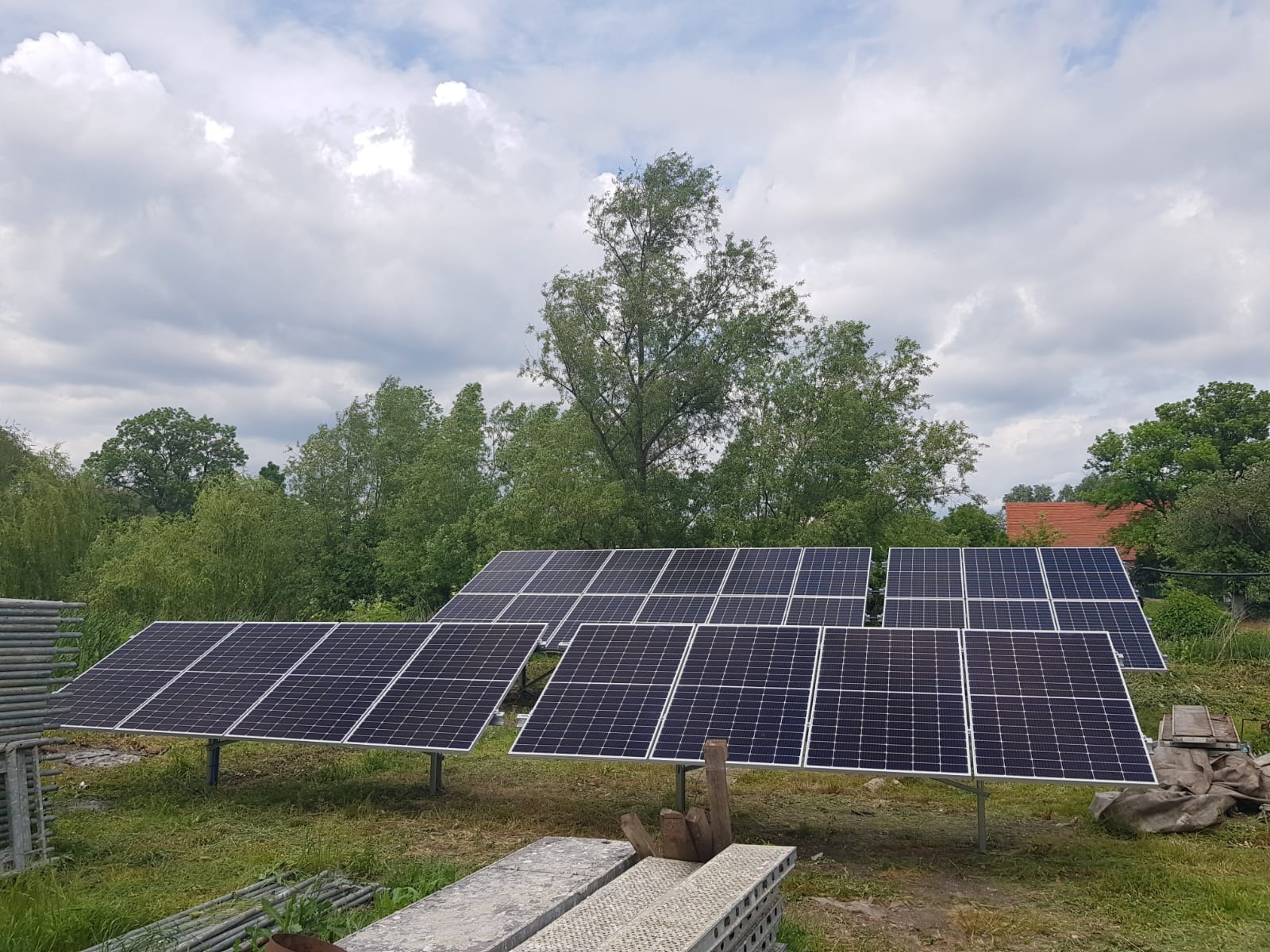 Rząd paneli słonecznych ustawionych na trawiastym terenie, widoczne drzewa w tle i pochmurne niebo, instalacja w trakcie realizacji, elementy rusztowania w lewym dolnym rogu.