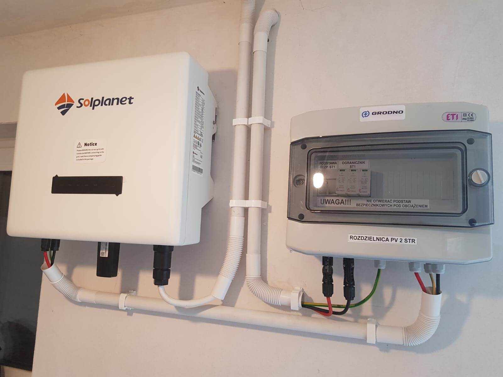 Zamontowany inwerter Solplanet i rozdzielnica PV 2 STR firmy Grodno na białej ścianie, z widocznymi kablami i rurami ochronnymi. Elementy instalacji fotowoltaicznej wewnątrz budynku.
