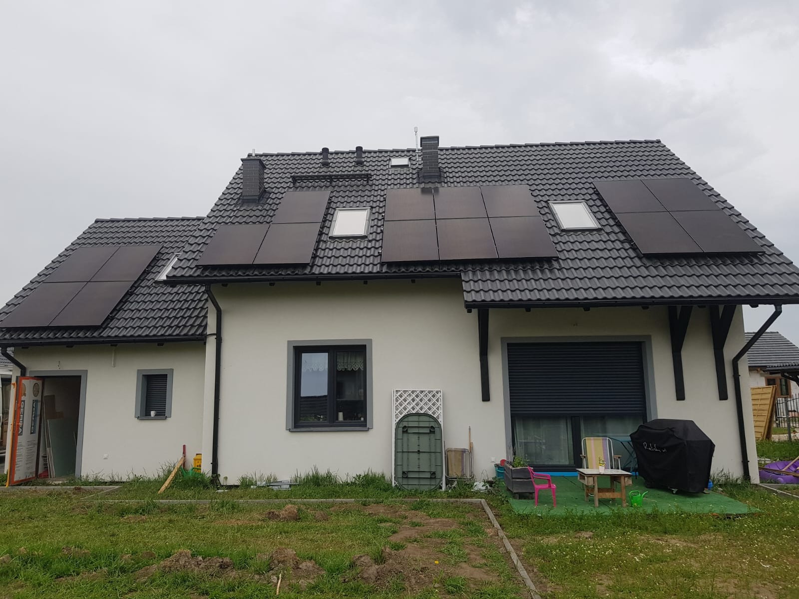 Dom jednorodzinny z zainstalowanymi panelami słonecznymi na dachu pokrytym szarą dachówką. Na dachu widoczne okna dachowe. Na trawniku przed domem widoczne ślady prac ziemnych.