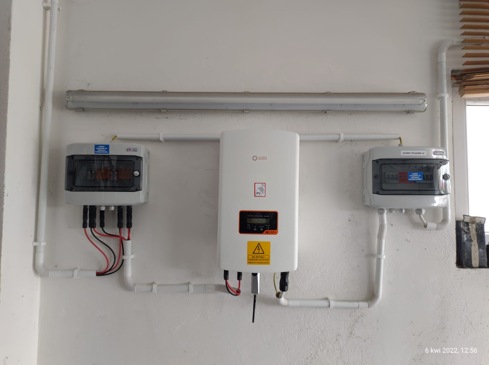 Biały inwerter fotowoltaiczny marki Solis zamontowany na białej ścianie, z widocznymi przewodami i skrzynkami rozdzielczymi po bokach, oraz rurami instalacyjnymi z tworzywa sztucznego.