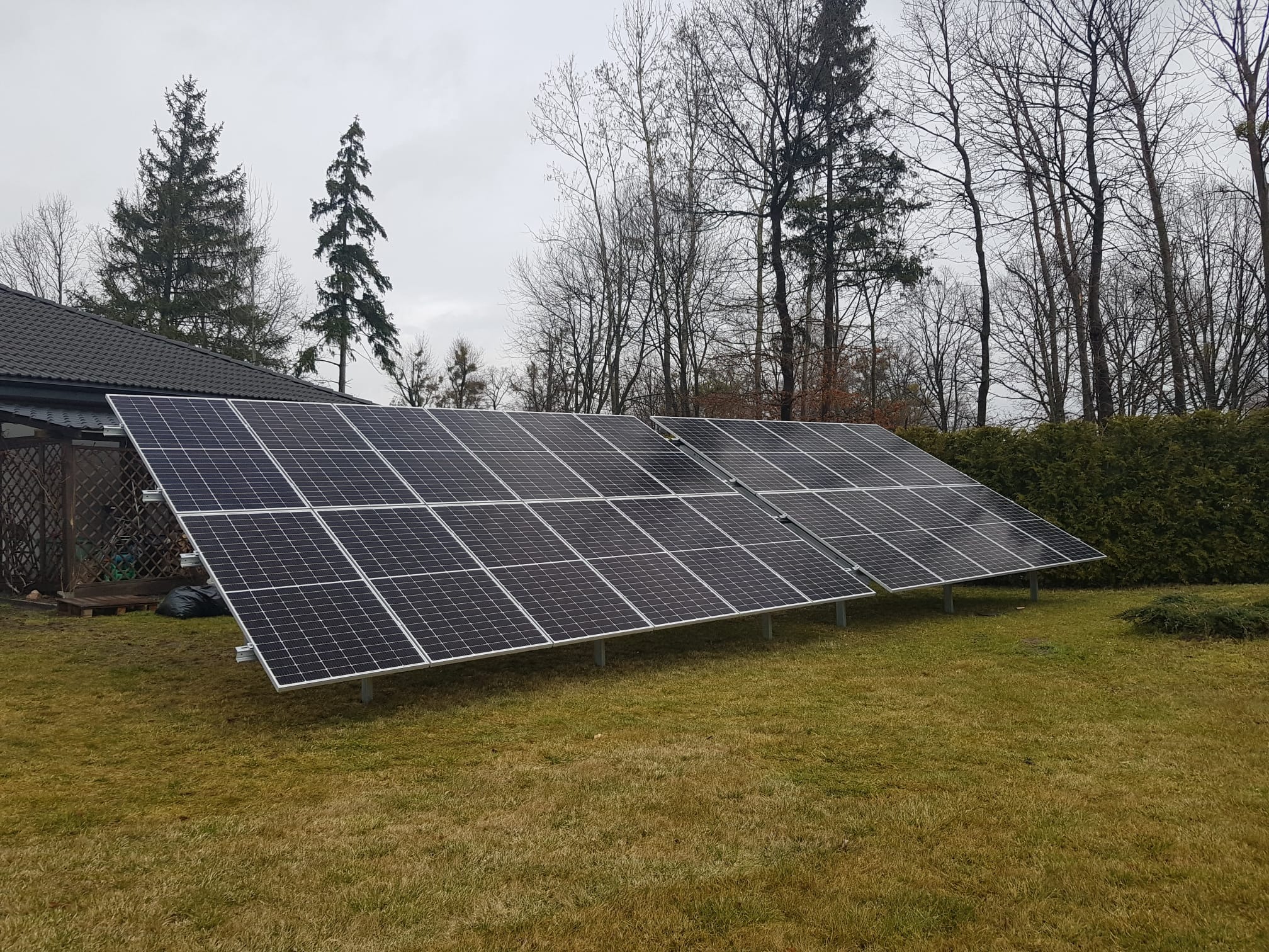 Instalacja fotowoltaiczna na trawniku przed domem jednorodzinnym, widoczne panele słoneczne ustawione pod kątem na metalowych wspornikach, w tle drzewa i krzewy.