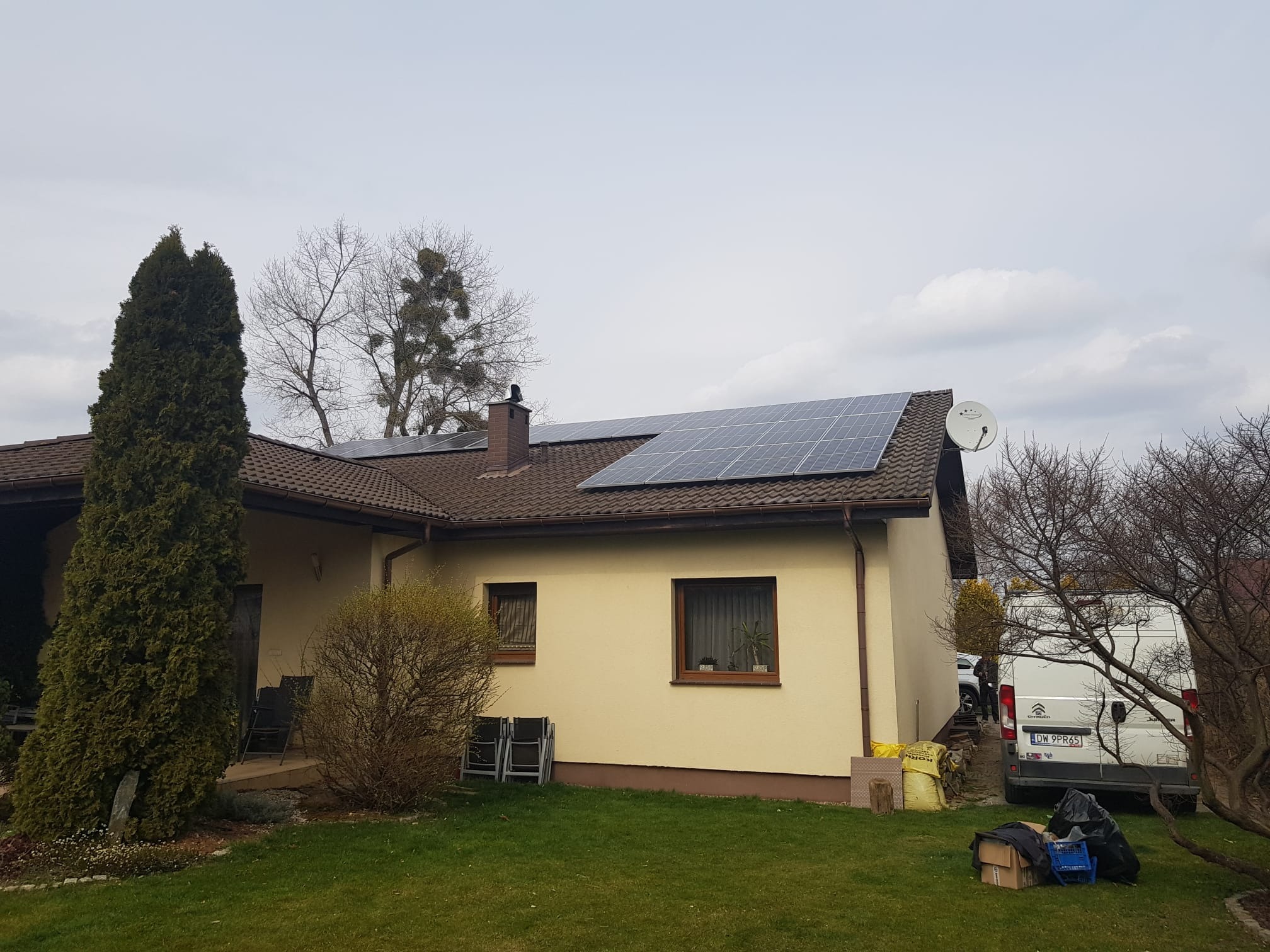 Panele słoneczne na brązowym dachu domu jednorodzinnego z kremową elewacją, widoczny samochód dostawczy firmy instalacyjnej i zadbany trawnik.
