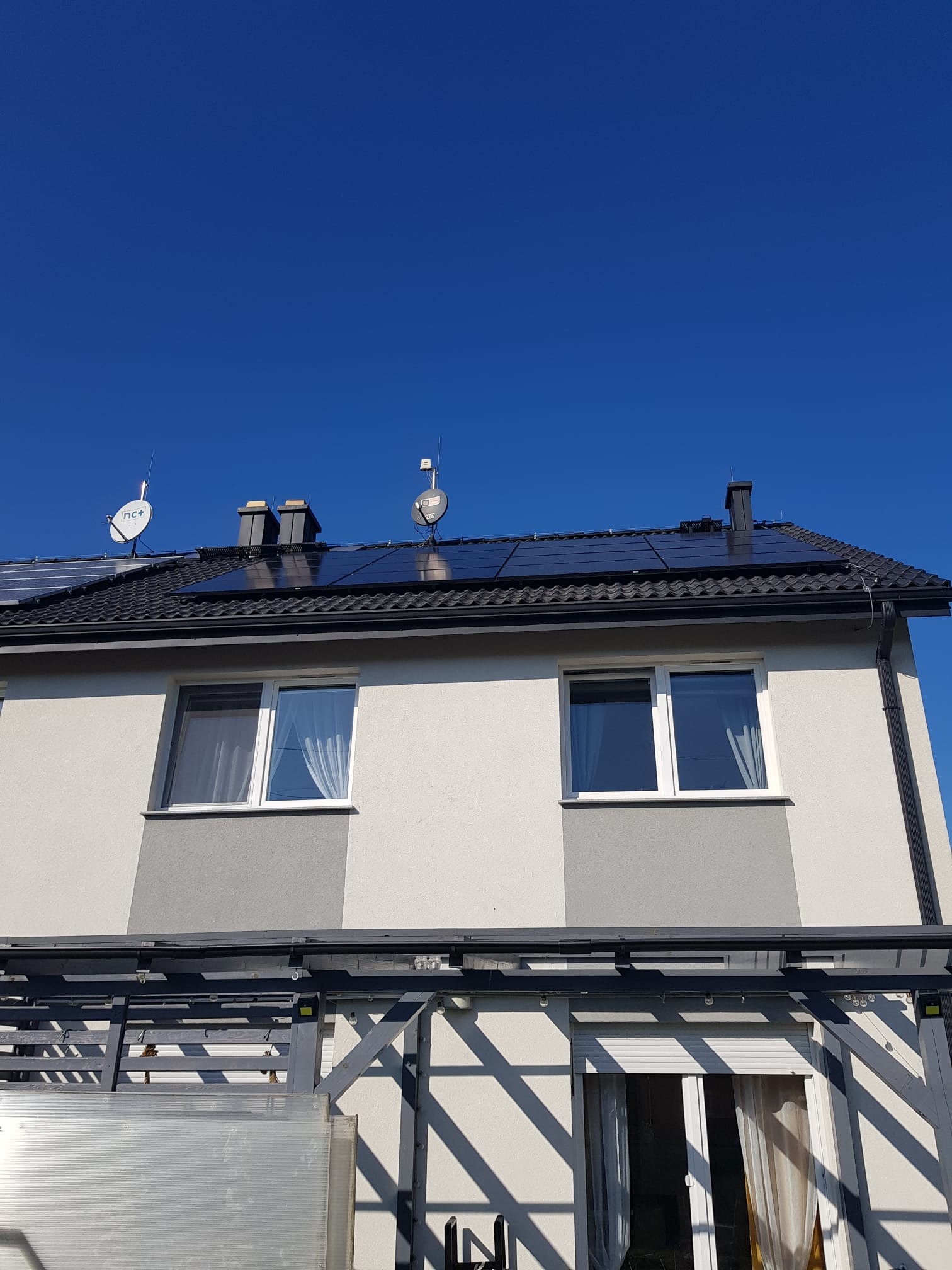 Panele słoneczne na dachu dwukondygnacyjnego domu z antenami satelitarnymi, widoczne z dołu w słoneczny dzień.