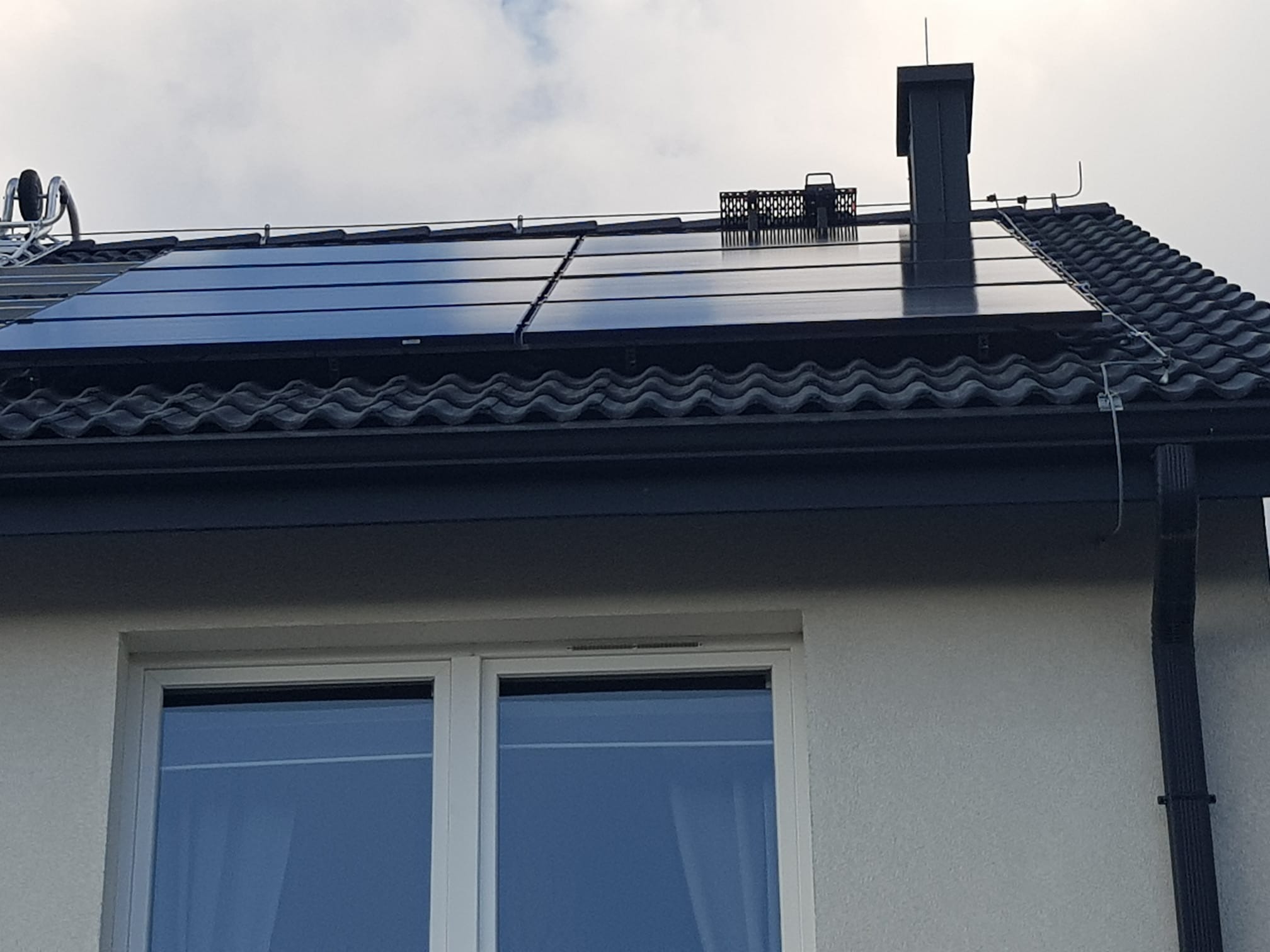 Czarne panele słoneczne na ciemnym, spadzistym dachu z dachówki ceramicznej, widoczne z dołu, z elementami mocującymi, odgromnikiem i kominem. W tle pochmurne niebo i fragment okna.