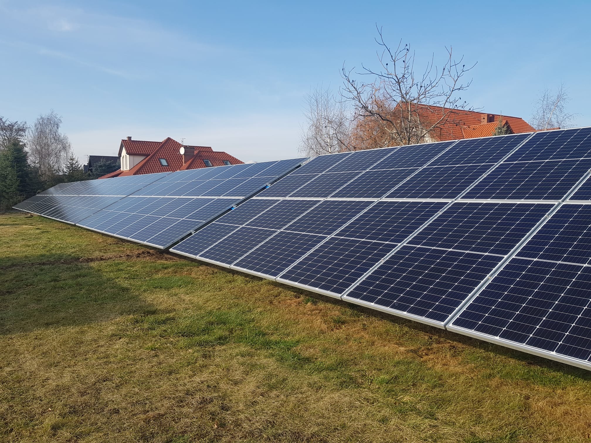 Długi rząd paneli fotowoltaicznych ustawionych na trawniku przed domem z czerwoną dachówką w słoneczny dzień.