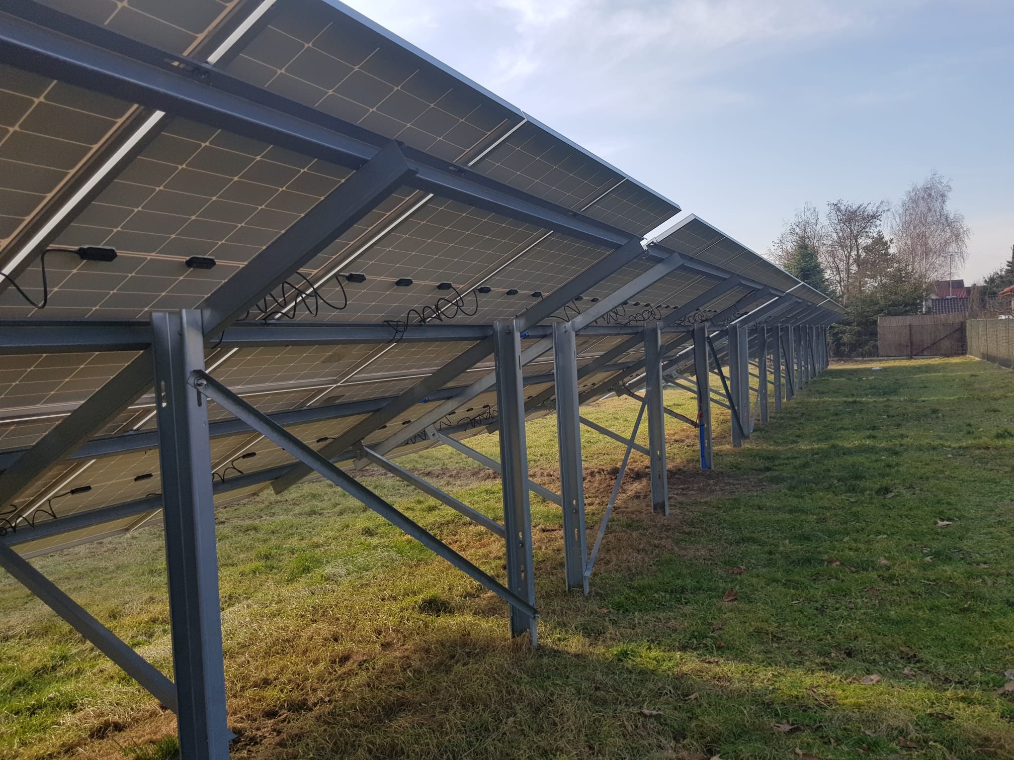Widok od spodu na panele słoneczne zamontowane na metalowej konstrukcji ustawionej na trawniku, z widocznymi przewodami i elementami mocującymi.